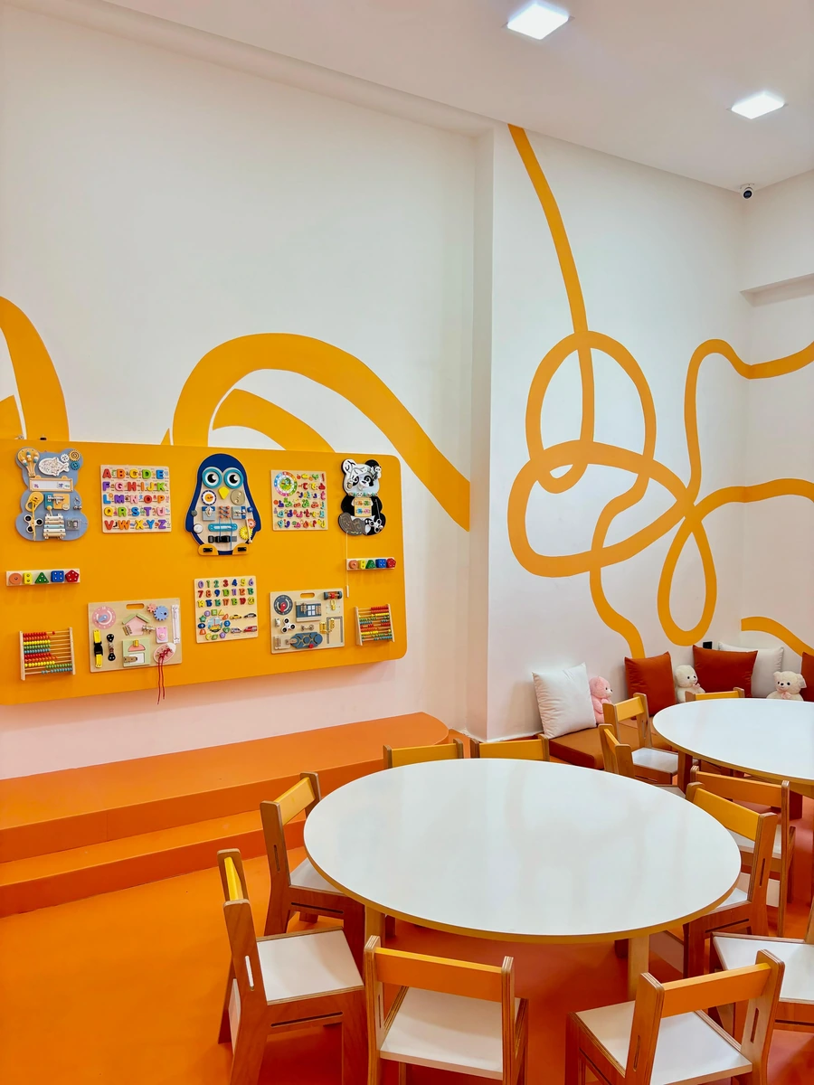 Salle Ptichou aux murs jaunes, avec panneaux de jeux, tables rondes et petites chaises.